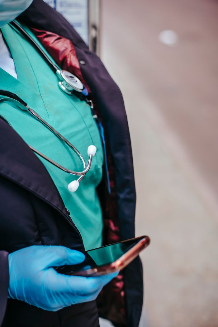 Crop anonymous female doctor in uniform with stethoscope touching black screen on cellphone in town